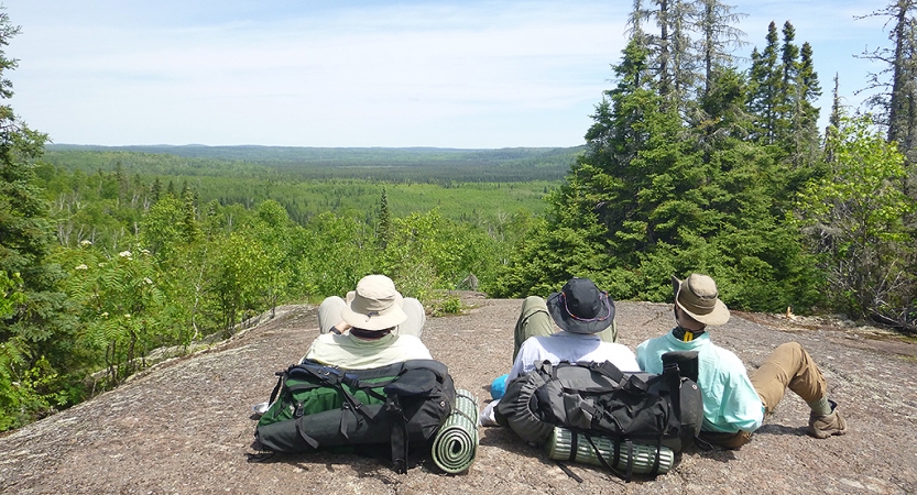 Three people sit on a rocky overlook and lean back on their backpacks, taking in the vast green forrest below.